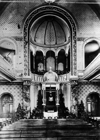 Black and white photograph of the interior of a synagogue, with Torah shrine, benches and circular gallery