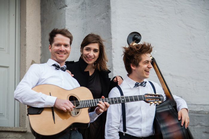 Gypsy-Swing und Klezmer mit der Band "Brise Manouche" Die Sängerin der Band "Brise Manouche" steht zwischen zwei Musikern der Band, welche ihre Instrumente in den Händen halten. Die drei Musiker sind lächelnd abgebildet und Wind weht sichtbar durch deren Harre.