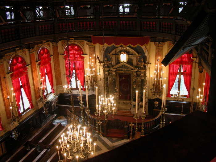 Geschmückter Innenraum der Synagoge Spagnola in Venedig mit Toraschrein, brennenden Kerzen an den Kronleuchtern und roten Fenstervorhängen 