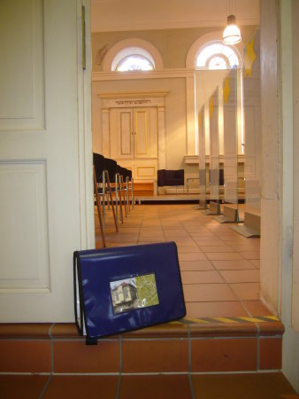 Ein blauer Rucksack steht am Eingang zum Betsaal der Kleinen Synagoge. Im Hintergrund sieht man den Toraschrein. Im Rucksack befinden sich die Rätselmaterialien für das Detektivspiel.