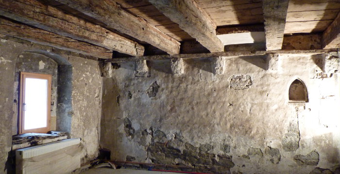Ansicht eines mittelalterlichen Raumes mit Steinwand mit spitzbogiger Nische für eine Lichtquelle und bemalter Holzbalkendecke.
