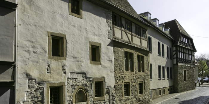Eine mit Kopfstein gepflasterte Straße im Sonnenlicht. Eine Reihe unterschiedlich alter Häuser ist zu sehen, eines mit Fachwerk, eines aus grob verputztem Mauerwerk, ein zeitgenössisches Gebäude.