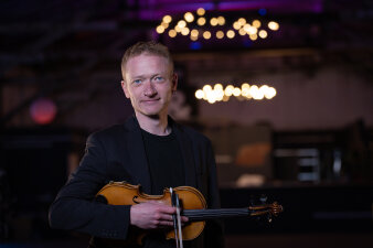 ein Mann mit Violine unter dem Arm steht in einem dezent beleuchteten Raum