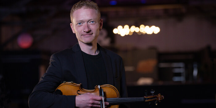 ein Mann mit Violine unter dem Arm steht in einem dezent beleuchteten Raum