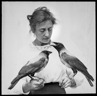 auf den beiden Händen einer Frau sitzt jeweils ein rabenartiger Vogel