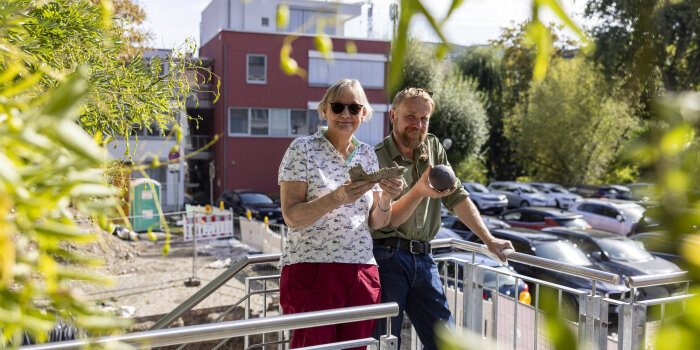 eine Frau und ein Mann stehen an einem Geländer und halten je einen Gegenstand in der Hand