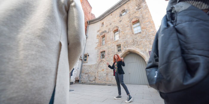vor einem historischen Gebäude steht eine Frau mit langen roten Haaren und spricht