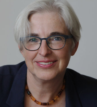 Eine Frau mit kurzen grauen Haaren und schwarz umrandeter Brille lächelt in die Kamera. Sie trägt eine Kette aus verschiedenfarbigen Bernsteinperlen und ein dunkelblaues Jackett.