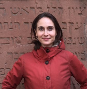 Eine Frau mit dunklen Haaren in rotem Anorak steht vor einer rötlich-braunen Steintafel, auf der hebräische Schriftzeichen eingraviert sind, und blickt in die Kamera.