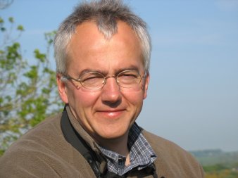 Ein Mann mit silberner Nickelbrille und kurzen grauen Haaren lächelt mit braun gebranntem Gesicht in die Kamera. Er steht im Sonnenschein, im Hintergrund ist blauer Himmel zu sehen. Am linken Bildrand sind grüne Zweige zu sehen, am rechten unteren Bildrand erkennt man in der Ferne eine grüne hügelige Landschaft.