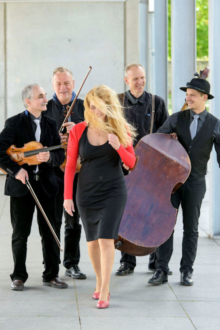 Eine Frau, deren Haare im Wind wehen, schreitet voran. Im Hintergrund stehen vier Männer der Band mit Instrumenten in den Händen. 