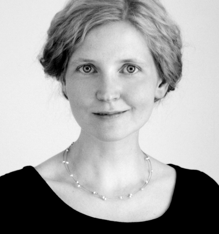 Klassisches Schwarz-Weiß-Porträtfoto: Eine Frau mit leicht gelockten, hinten zusammengefaßten hellen Haaren schaut gerade in die Kamera. Sie trägt eine Perlenkette und einen schwarzen Pullover.