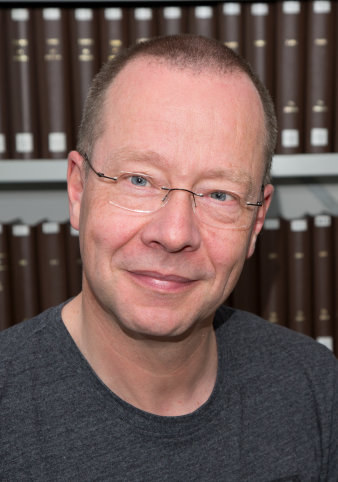 Ein Mann mit kurzen Haaren, einer randlosen Brille und einem dunkelgrauen T-Shirt blickt freundlich in die Kamera. Im Hintergrund sind Bücherregale zu erkennen.
