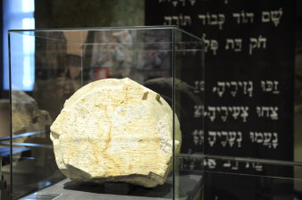 Vitrine mit einem bearbeiteten Stein im Vodergrund, dahinter Fahne mit hebräischer Inschrift