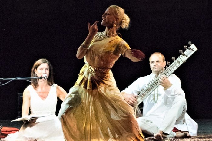tanzende Frau im goldenen Kleid, dahinter zwei Musiker/innen mit Zupfinstrument und Mikrofon