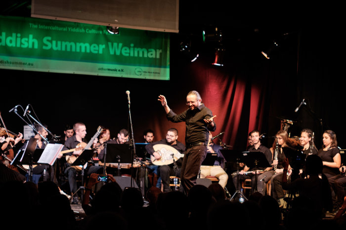 Bühne mit schwarz gekleideten Musikern mit unterschiedlichen Instrumenten, davor Sänger, Publikum von hinten
