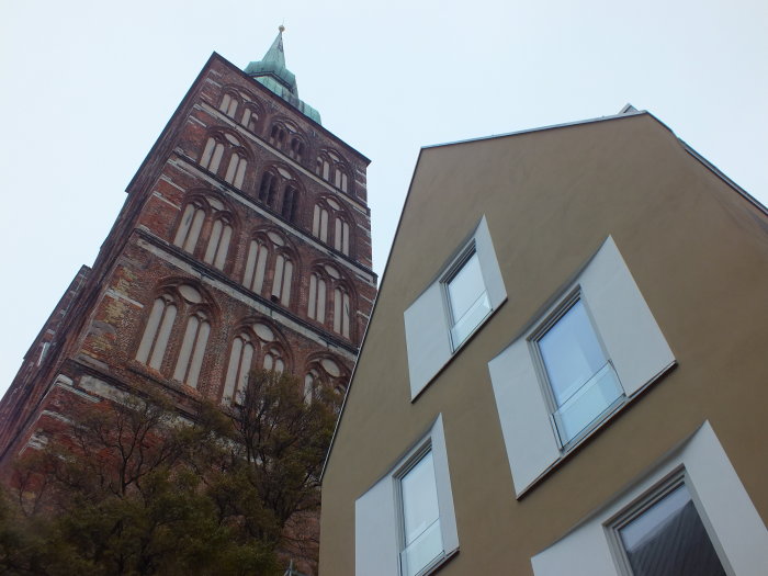 Links im Bild und im Hintergrund ragt ein gotischer Backsteinkirchturm in den Himmel. Auf der rechten Bildhälfte und eher im Vordergrund der erste Stock und das Dachgeschoss eines modernen, beige verputzten Wohnhauses