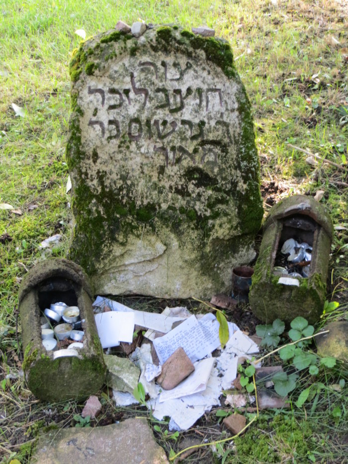 Das Foto zeigt einen verwitterten und bemoosten Grabstein mit hebräischen Schriftzeichen. Auf ihm liegen nach jüdischer Tradition kleine Steine zur Erinnerung an den Verstorbenen. Vor dem Stein sind Papierzettel mit Nachrichten an den Verstorbenen gestapelt. 