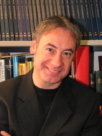 Ein Mann in einem schwarzen Sakko steht vor einem Bücherregal und blickt in die Kamera. 