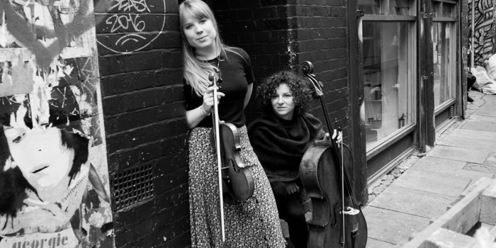 Eine Frau mit Violine und eine Frau mit Cello stehen an einer Häuserwand. 