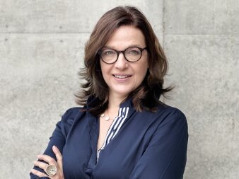 Portrait einer Frau im blauen Oberteil und mit Brille vor einer hellen Steinwand