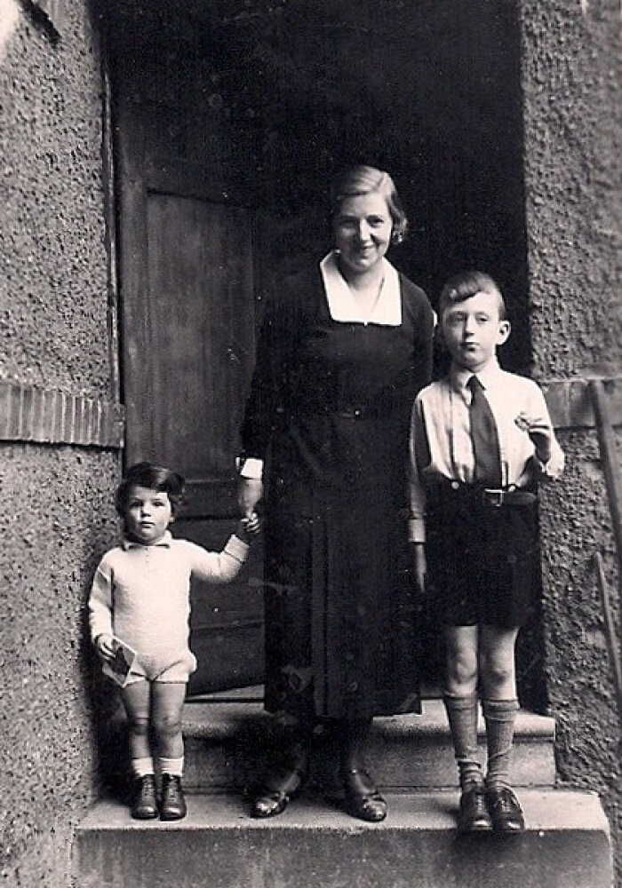 Schwarz-weiß Fotografie einer Frau in einem langen Kleid gemeinsam mit ihren beiden Kindern vor einem Hauseingang