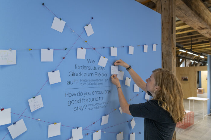 Mitmachangebot zur Ausstellung - Galerie Waidspeicher Eine junge Frau heftet einen Notizzettel an eine blaue Wand