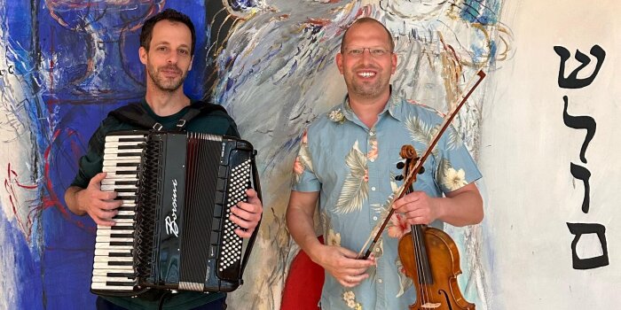 Zwei Männer mittleren Alters halten als Instrumente ein Akordeon und eine Violine in den Händen