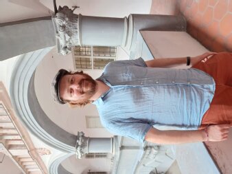 Mann mittleren Alters mit roten Haaren, Mütze und helblauem Hemd vor zwei Säulen stehend