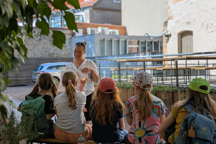 Fünf Kinder sitzen vor der Alten Synagoge und lauschen den Worten einer Pädagogin