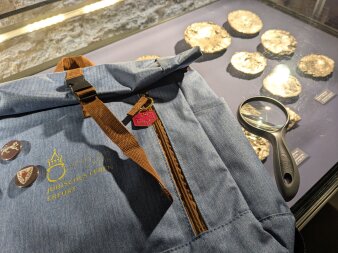 Foto: Detektivtour mit Rucksack in der Alten Synagoge © Stadtverwaltung Erfurt Ein kleiner, graublauer Rucksack mit Lupe und Buttons liegt auf einer Museumsvitrine mit gold-glänzenden Exponaten