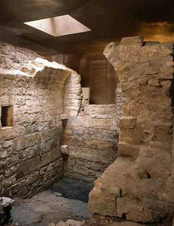 Mauerreste der mittelalterlichen Mikwe. Reste eines kleinen Raumes mit Tonnengewölbe, umgeben von Ausschnitten des bronzefarbenen Schutzbaus.
