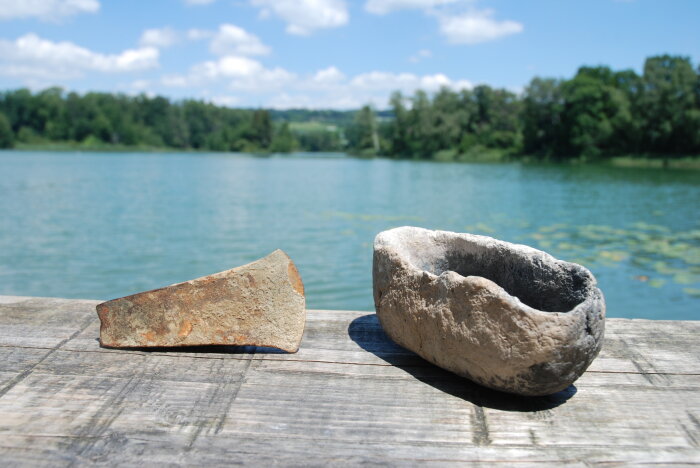 Zwei versteinerte Gegenstände auf einem Holzsteg, dahinter ein See 