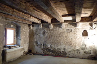 Ansicht eines mittelalterlichen Raumes mit Steinwand mit spitzbogiger Nische für eine Lichtquelle und bemalter Holzbalkendecke.