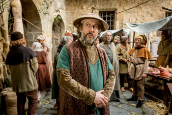 Foto: MDR-Film „Der Schatz von Erfurt" 2016 © MDR / Andreas Lander Das Foto ist eine Standaufnahme von Dreharbeiten zum Film „Die Pest, die Angst und der Schatz von Erfurt“, in dem es um den Schatz des Kalman von Wiehe geht. Er war ein jüdischer Kaufmann in Erfurt und kam, wie ein Großteil der jüdischen Gemeinde, im März 1349 bei einem Pogrom um. Das Bild zeigt einen Mann im zeitgenössischen Kostüm mit Judenhut und kurzem grauem Vollbart, der besorgt an der Kamera vorbei blickt. Im Hintergrund sind weitere mittelalterlich gekleidete Menschen vor Markständen auf mit Stroh bedecktem Steinboden zu sehen.