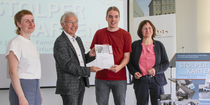zwei Frauen und zwei Männer stehen nebeneinander, die beiden Männer in der Mitte halten ein bedrucktes Blatt Papier