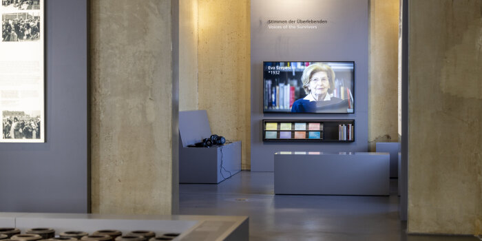 Ausstellungsraum, auf einem Bildschirm mit Sitzecke ist eine Frau zu sehen